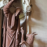 A statue, set into a wall, of a man and a goat. The goat is on its hind legs and a sugar lump has been balanced on its nose.