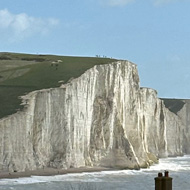 A view of the white cliffs which make up the Seven Sisters from the Seaford end with a train and houses in the foreground. Cliffs and sea going into the distance with mostly blue skies.