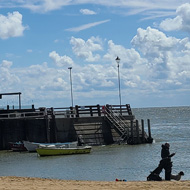 A bright Spring day. Lots of different white clouds against a blue sky, over a the sea and beach. There is a jetty with a large building on it.