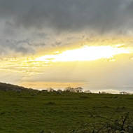 A lush green field sits under a densely clouded blue-grey sky, as the sun begins to descend in the distance