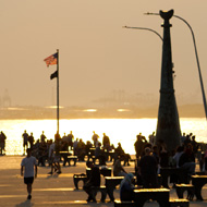 The Bay Ridge Pier in Brooklyn can be seen through a freeway underpass. Late afternoon light is gold and yellow tinted, and is reflecting sharply off an old sedan, and the white road markings. People on bikes and fishing can be seen on the pier itself in the background.