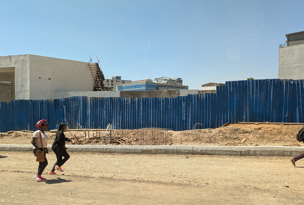 Two women walk in front of a tall blue corrugated fence which runs along a dusty roadside in the bright African sun behind it new buildings are being constructed