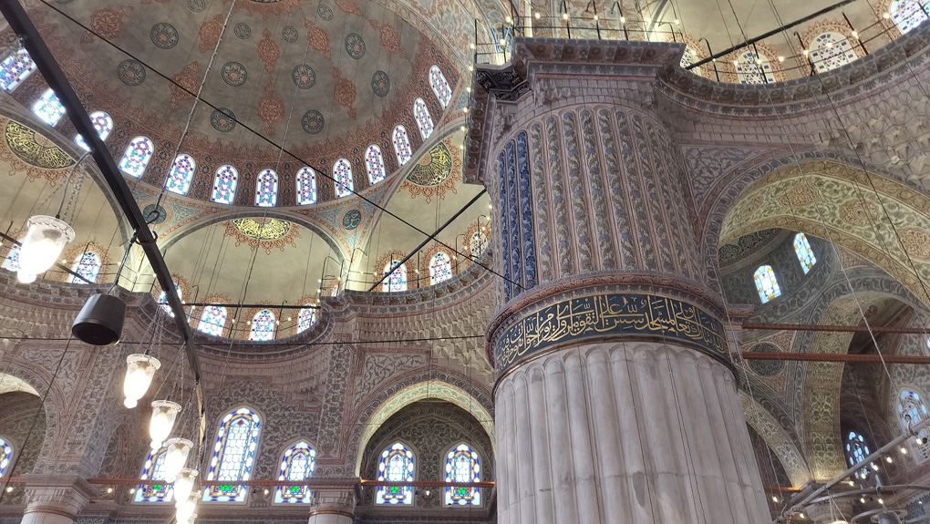 The interior of a mosque - looking upwards towards a dome, balconies and a pillar. The pillar has a tiled patteren on it, depicting rows of foliage - but three of the rows are a significantly deeper shade of blue.