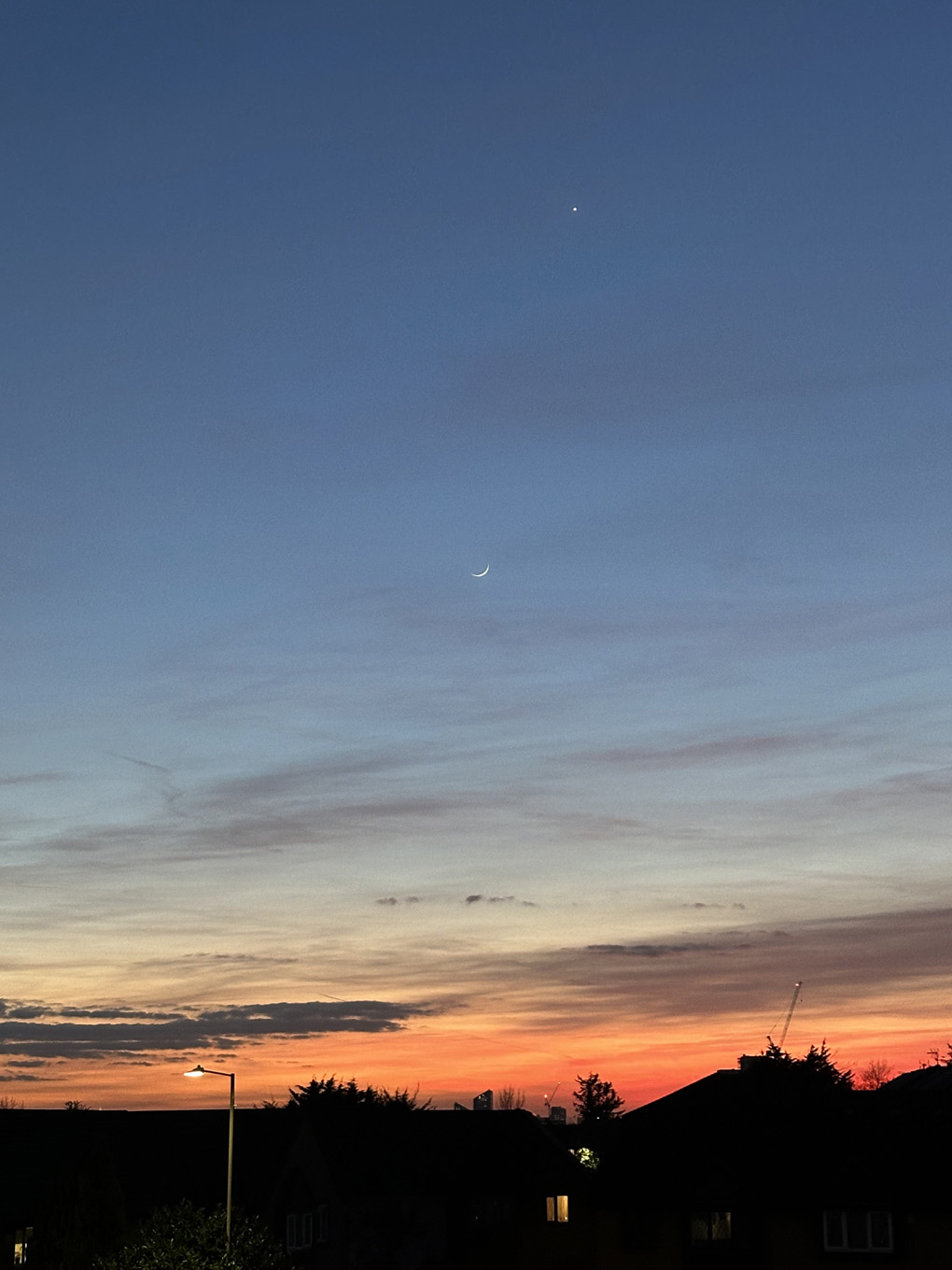 Red/orange sunset over rooftops in distance. Crescent moon and bright Venus in sky.