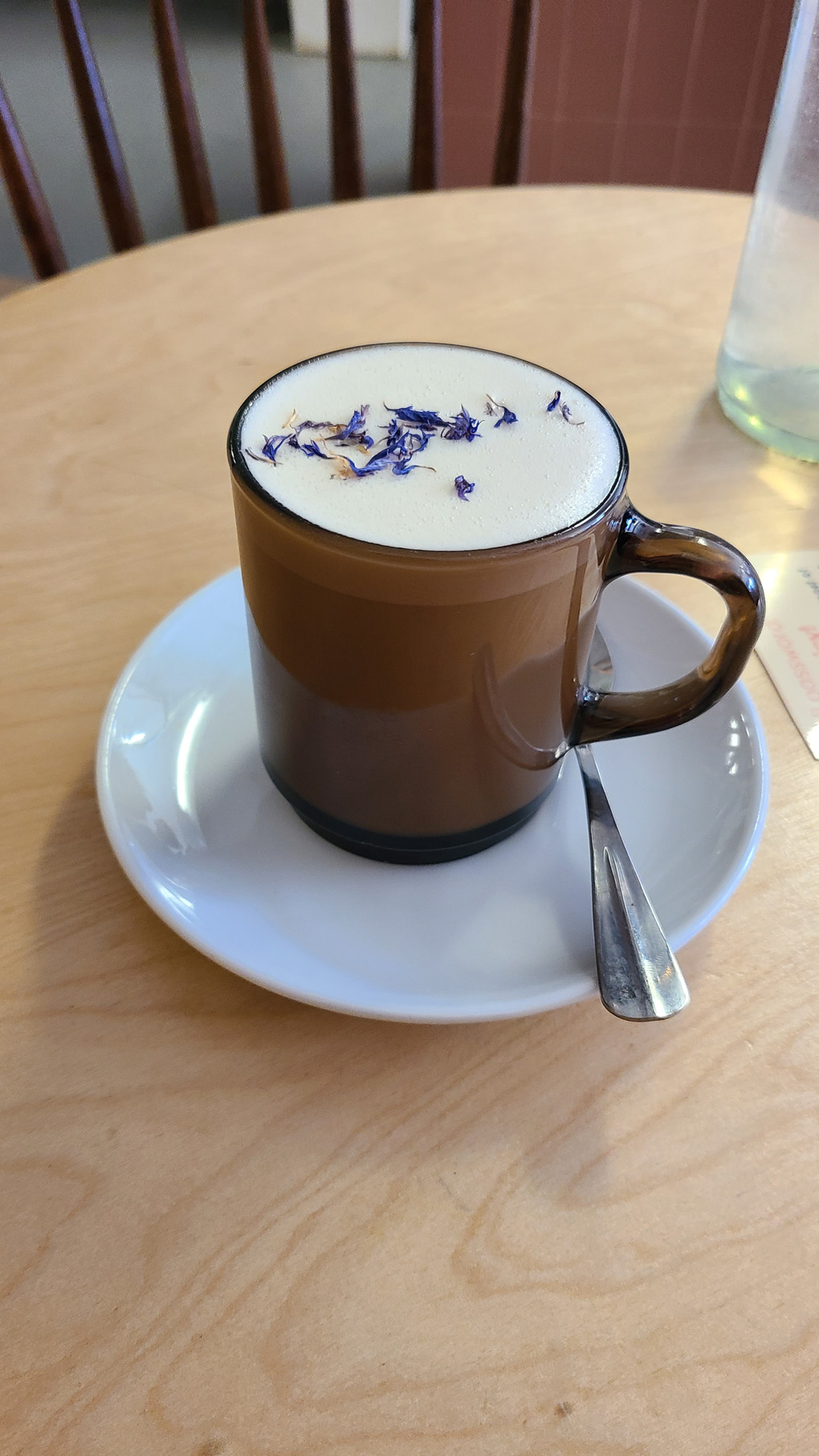 A brown glass mug on a white ceramic saucer. The mug is filled with creamy frothy liquid, topped with pretty purple and blue flower petals. The mugs handle is facing to the right, there is a silver teaspoon on the saucer under the handle. It's on a round, light wooden table.