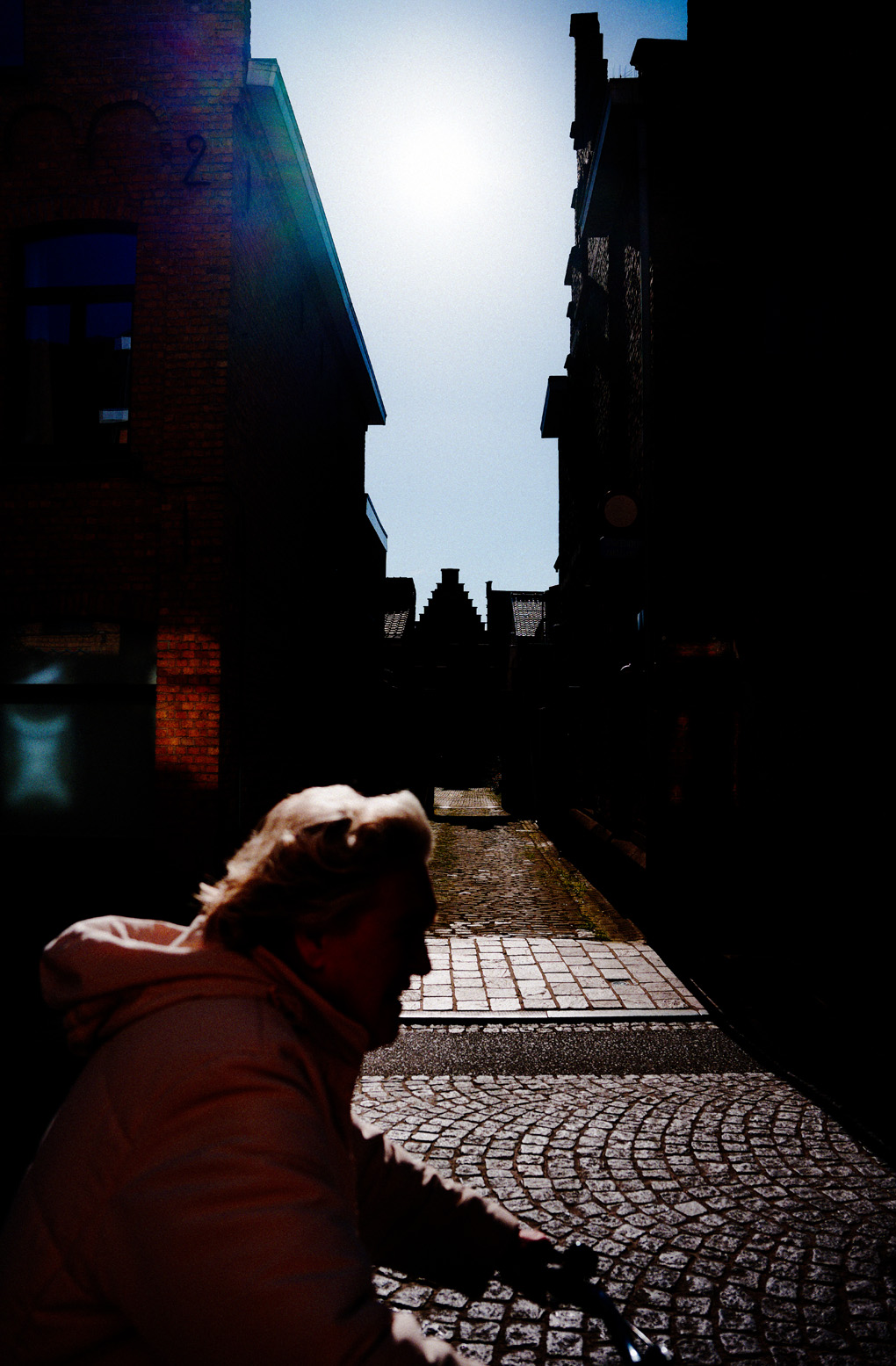 A woman rides a bicycle past a sunlit alley on a cobbled street in Ypres. Sunlight glances off her hair and jacket, and some geometric shapes in the background.