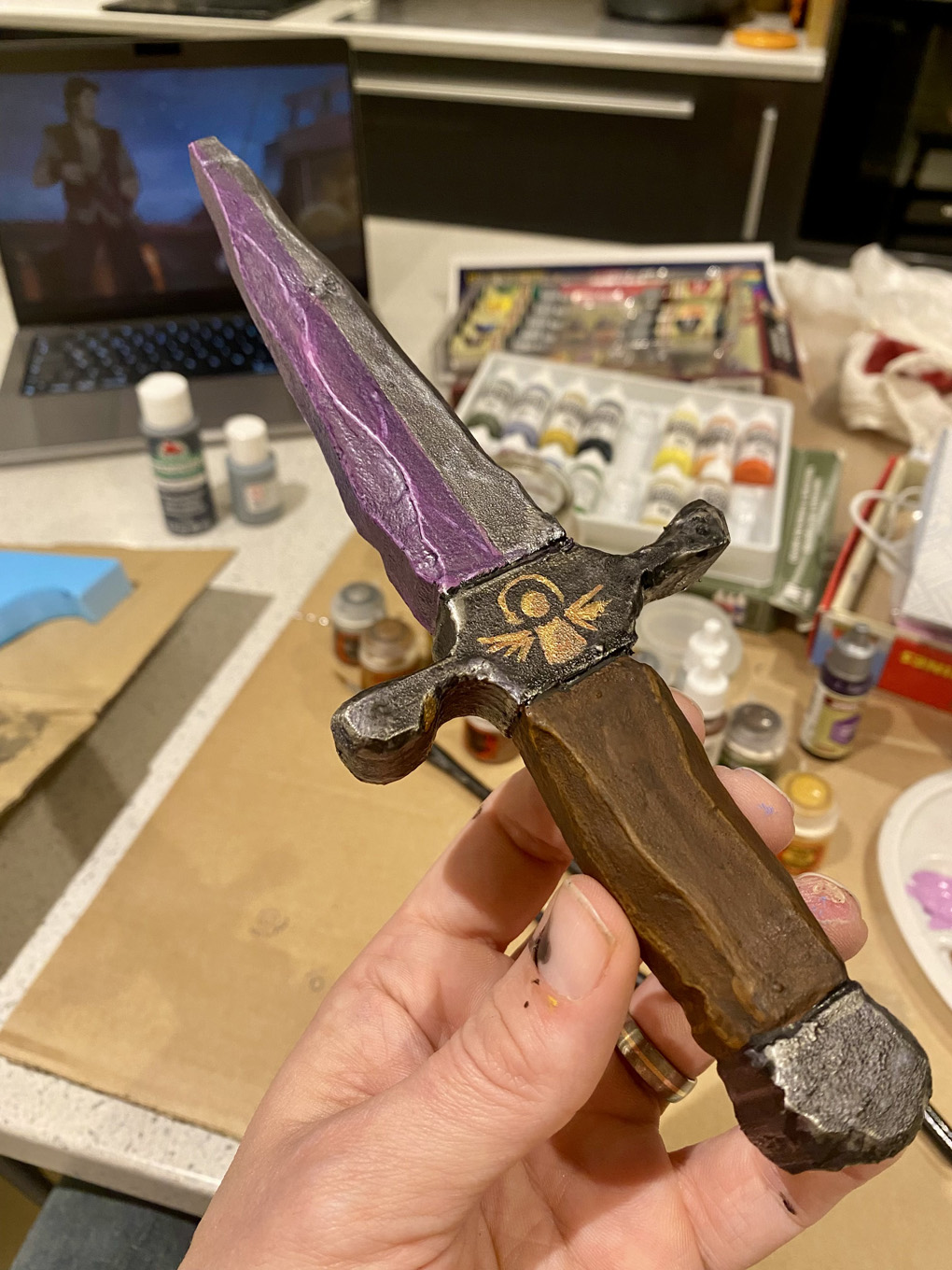 A model knife with a purple and silver blade, and a stylised angel on the hilt, being held in front of some model painting supplies.