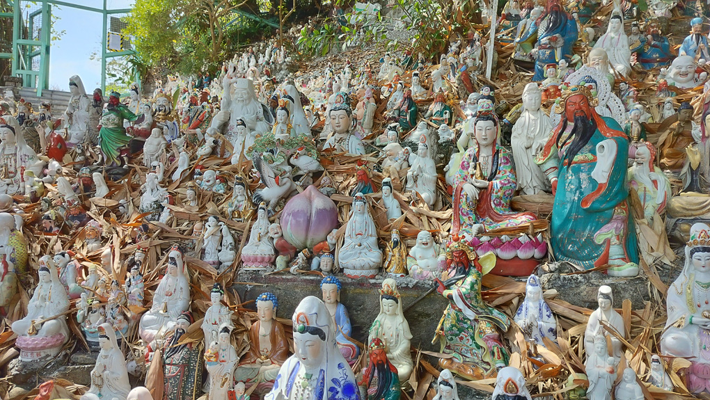 Along concrete steps, hundreds of ceramic models of Buddah and other Gods or religious deities are lined up. Leaves have fallen in amongst the statues, which gives a higgledy-piggledy view stretching up the steps.