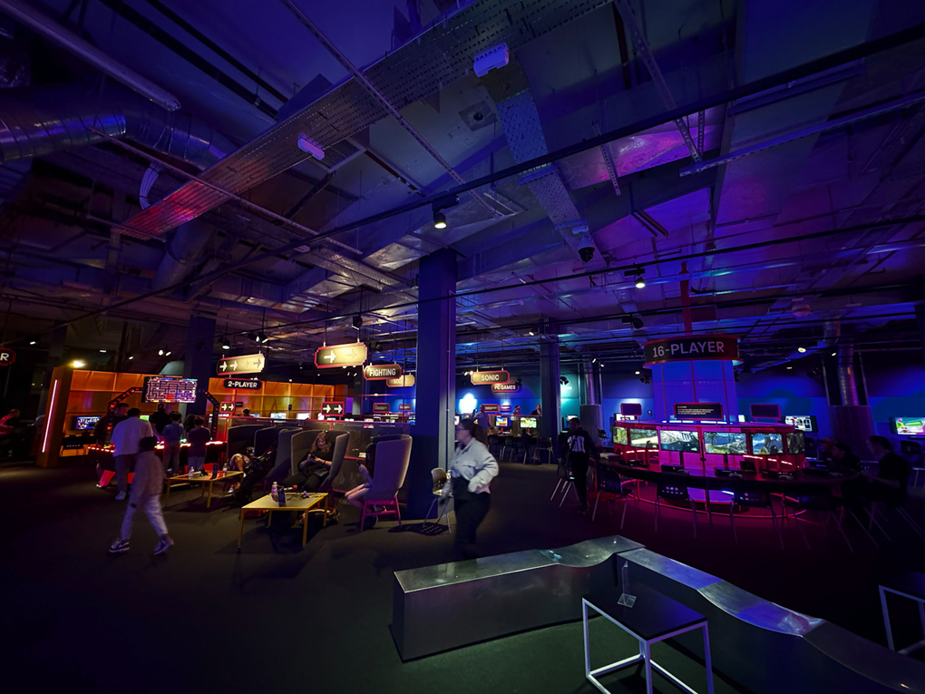 A dark underground room full of video game consoles and computers, sparsely lit by multi-coloured neon lighting