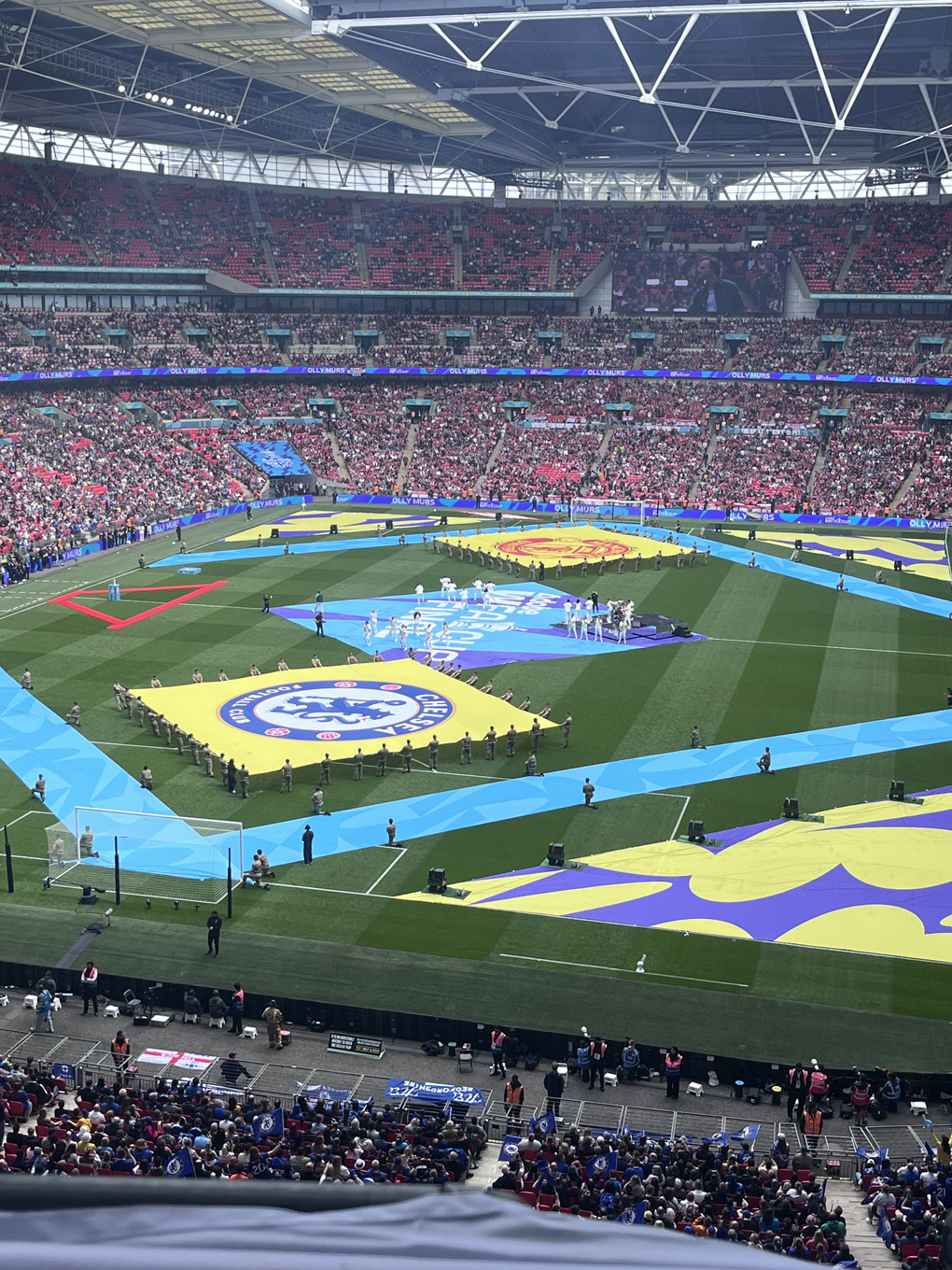 The football pitch at Wembley, with Chelsea and Manchester United team banners rolled out on the pitch, and the singer Olly Murs and dancers performing in the background
