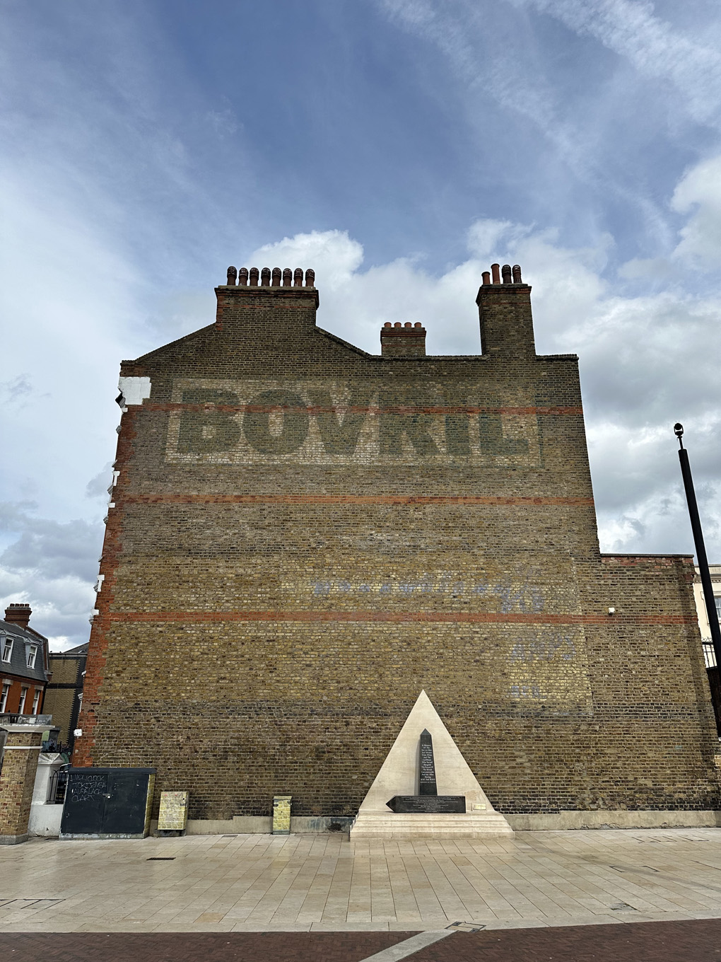 And old brick building with the large faded letters spelling BOVRIL near top. Pyramid and obelisk shaped war memorial at bottom. Blue sky and wisps of cloud.