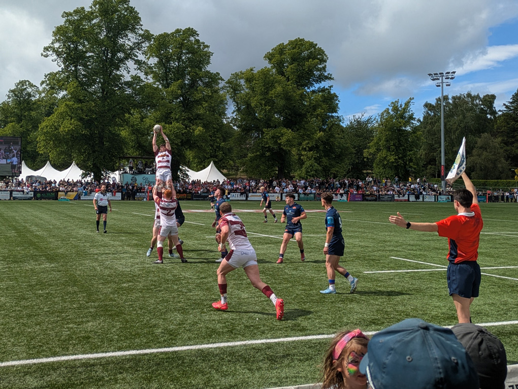 Rugby players leap at the line out in a game of sevens. in the background you can see the picturesque stadium lined by hundreds of fans framed by trees on a beautiful spring day