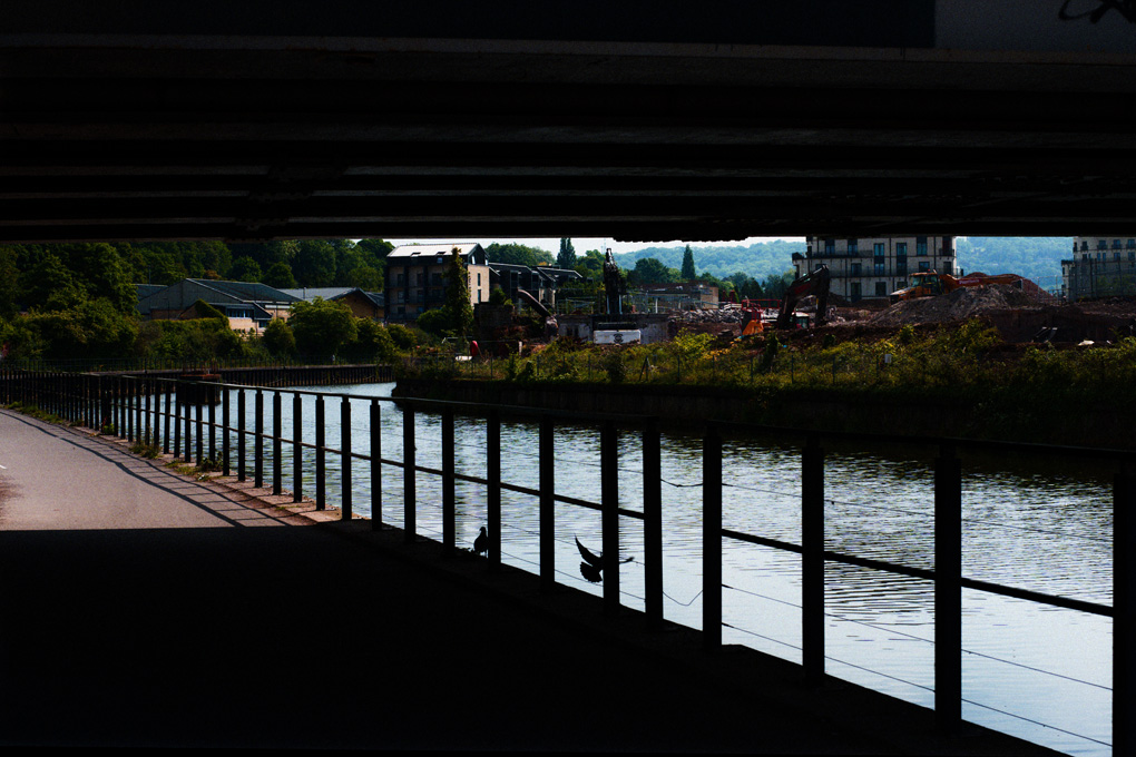 Two pigeons silhouetted behind the riverside railing on the Avon. One is in flight. In the distance the construction on the south side of the river is visible.