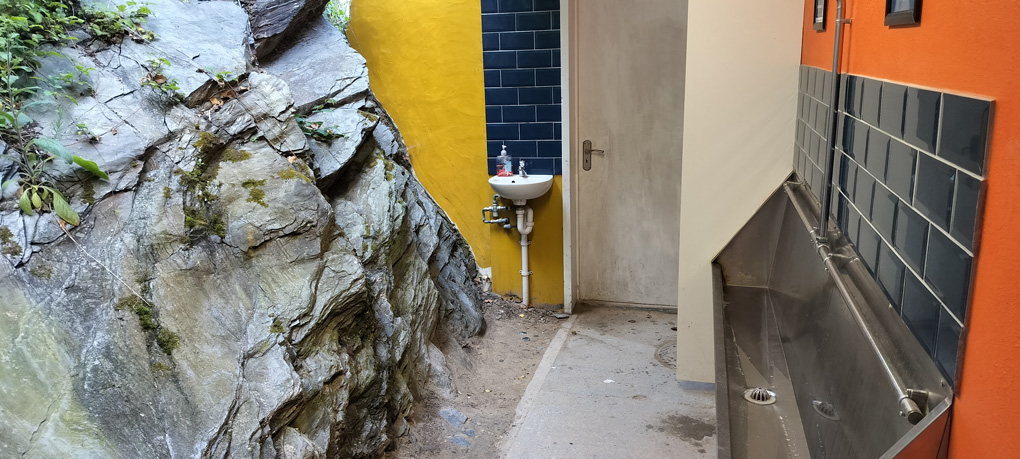 On the right of the image, a metal urinal in a men's toilet. In the centre the door and a sink. On the left hand side, an enormous rock formation with vines/leaves/moss which forms the left-hand wall.