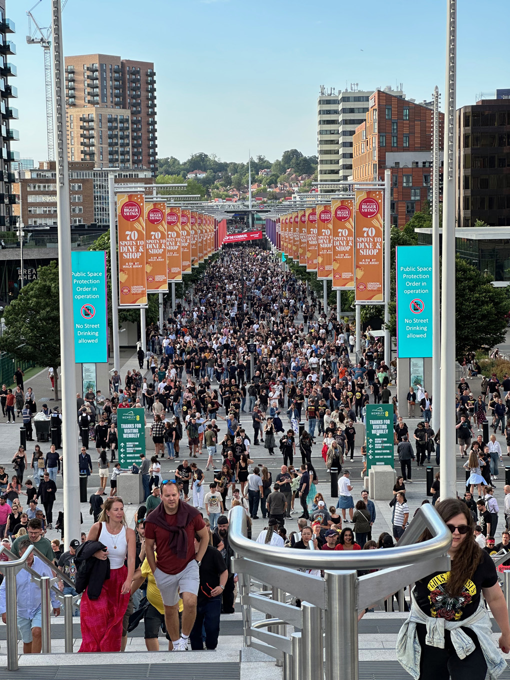 Crowd of people walking along big open path lined with signs and advertisements on either side. Wembley Stadium shot looking back towards station.