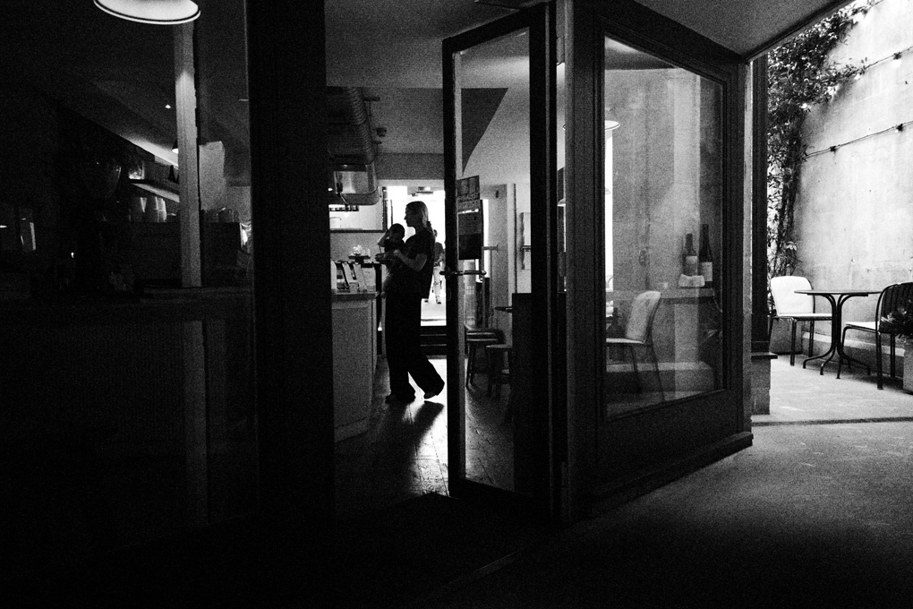 A woman holding her baby silhouetted in a coffee shop. Lit from behind, the natural shapes her body makes contrast against the straight lines and square angles of the doors, windows, and wall.