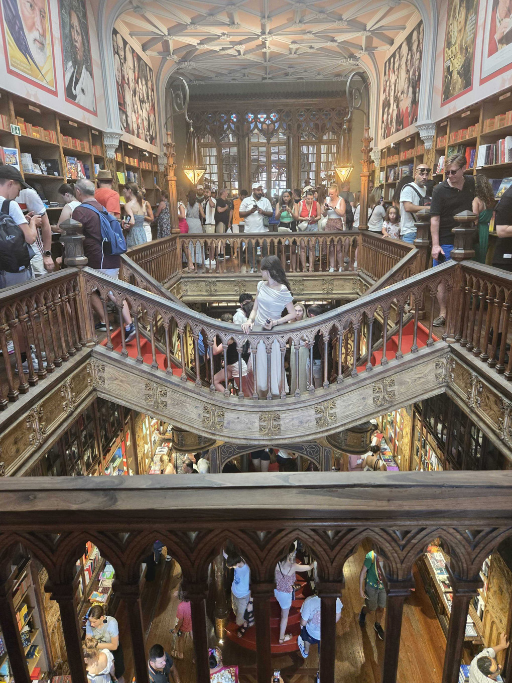 On the top floor of the bookshop, in the background are bookshelves, above the bookshelves are art deco style paintings decorating the walls. The majority of the room is taken up by the wooden frame of a grand staircase, in the centre is a bridge which leads to the steps. Through the wooden frame, you can see downstairs where the bottom of the red steps are visible along with more bookshelves. Both floors are full of people admiring the beauty and taking photos.