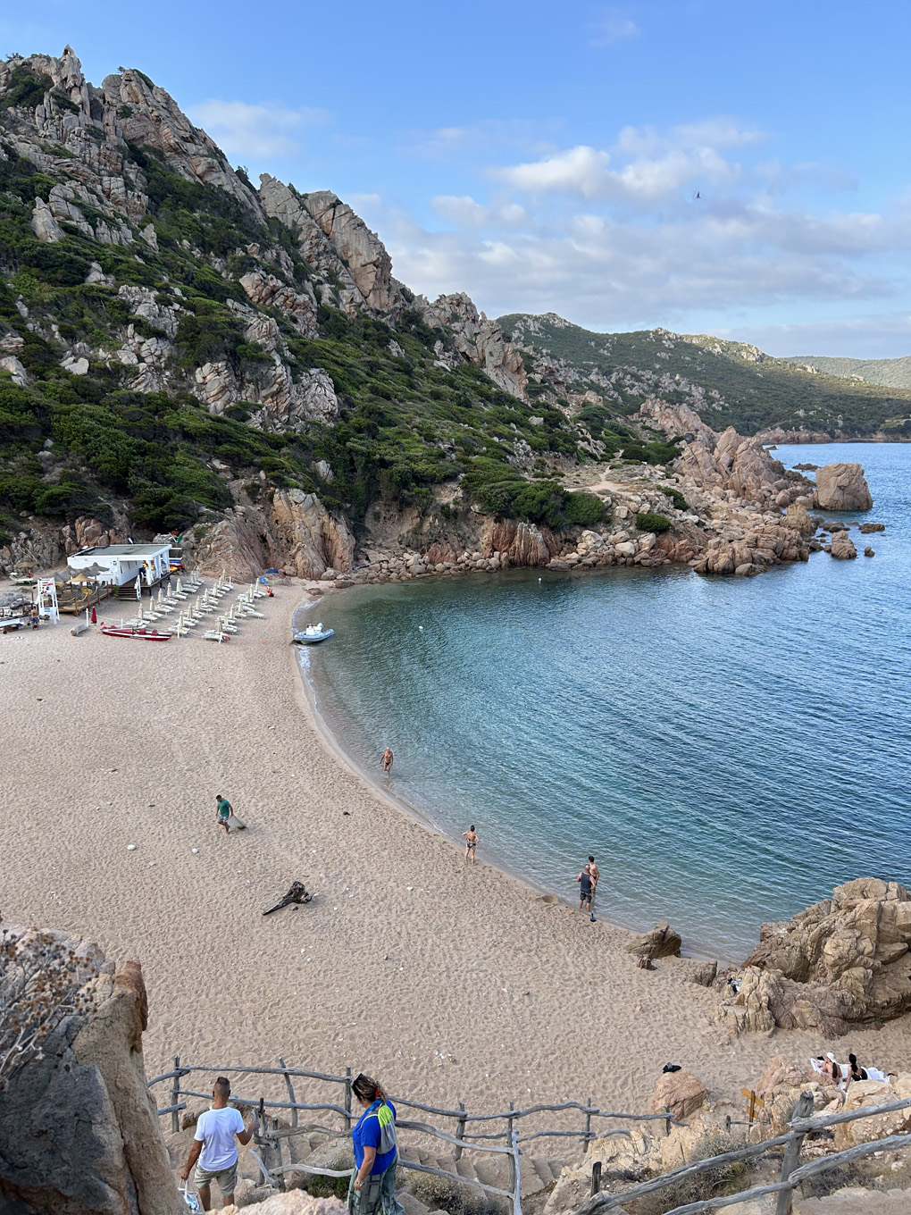 A scenic view of a small sandy beach nestled in a rocky cove with clear blue water. People are walking along the beach and swimming. Steep, rugged cliffs covered in greenery rise behind the beach, and a few beach chairs and umbrellas are visible on the left side near a white structure.