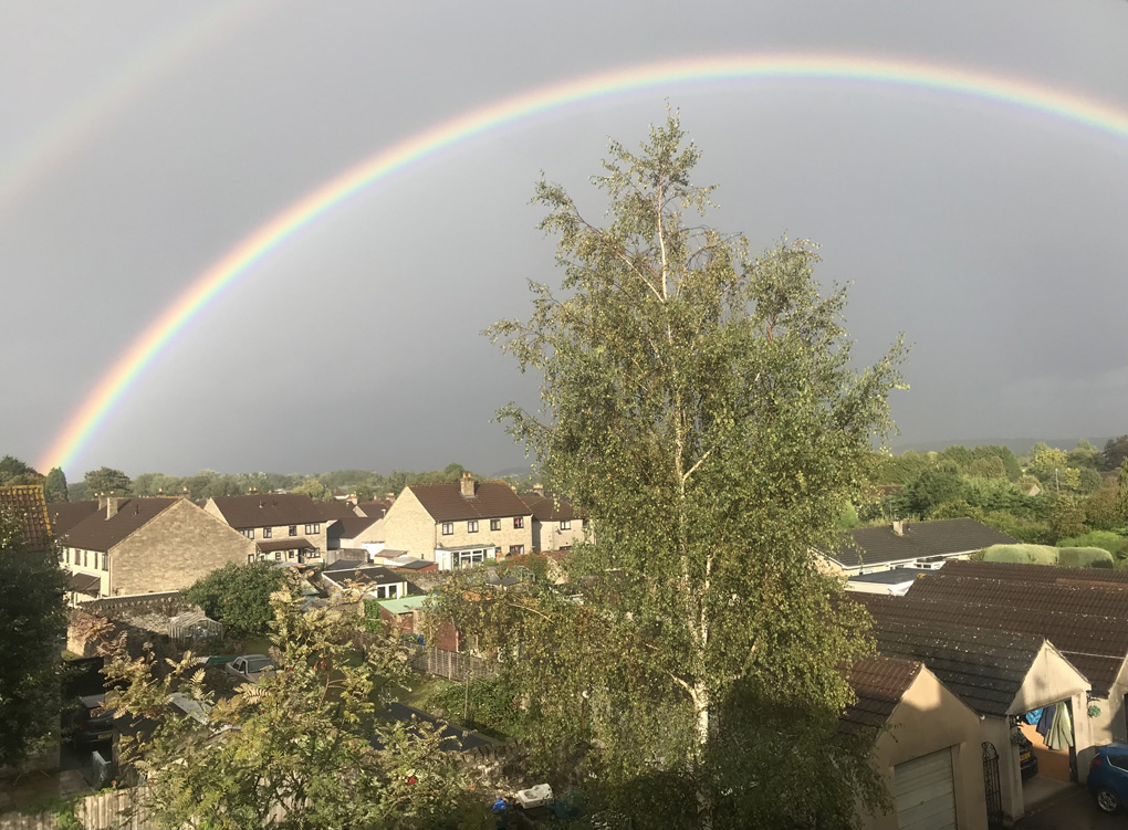 A bright and full semicircular colourful rainbow, stretched across a grey sky, above sun lit roofs and treetops.