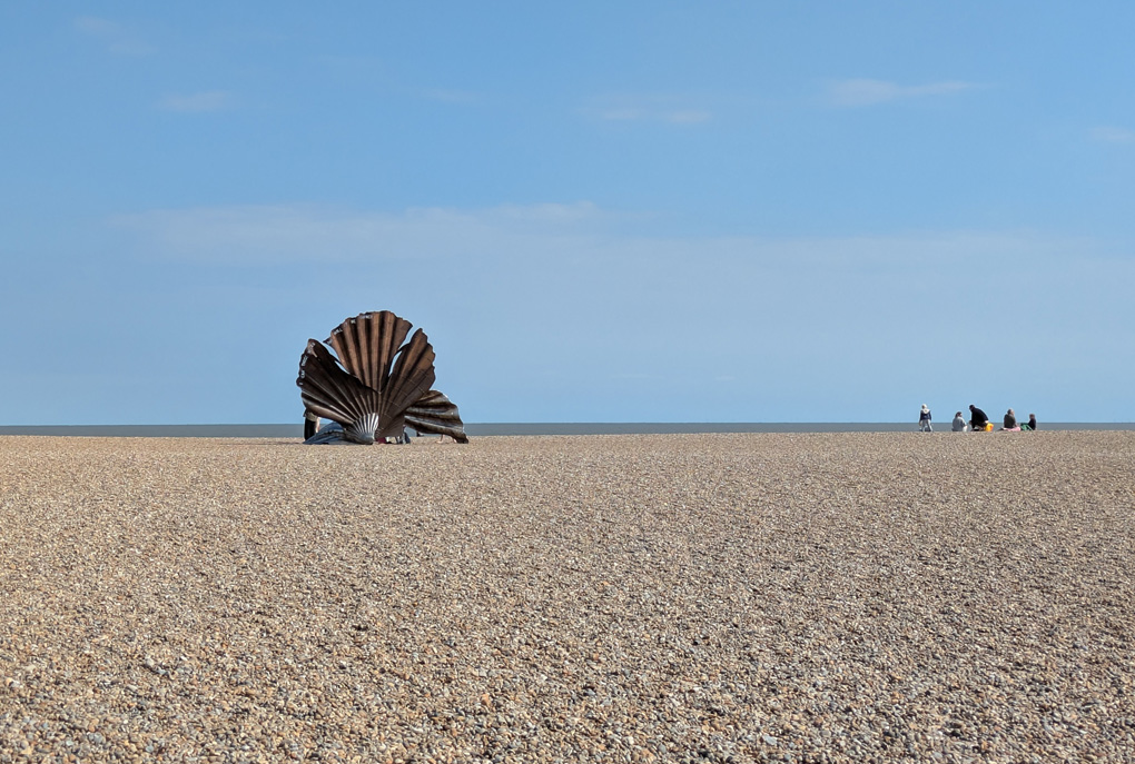 A wide pebble beach below a wide blue sky. On the beach a large sculpture of a scallop shell made from curving sheets of metal is set against the sky