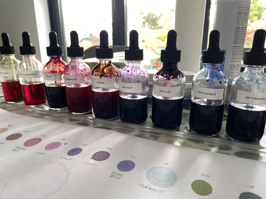 A line of labelled bottle droppers containing coloured inks, behind a piece of paper with drops of each colour.