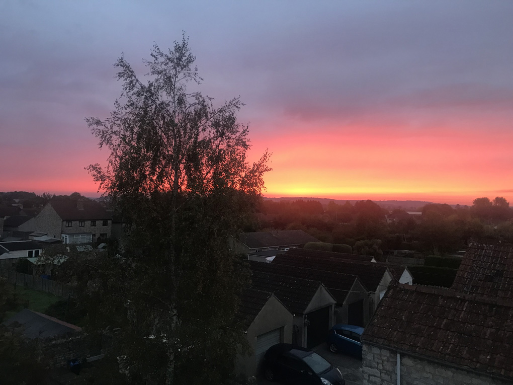 Bright orange ray of sun spearing over a dark horizon, eliminating rooftops and trees as is rises.