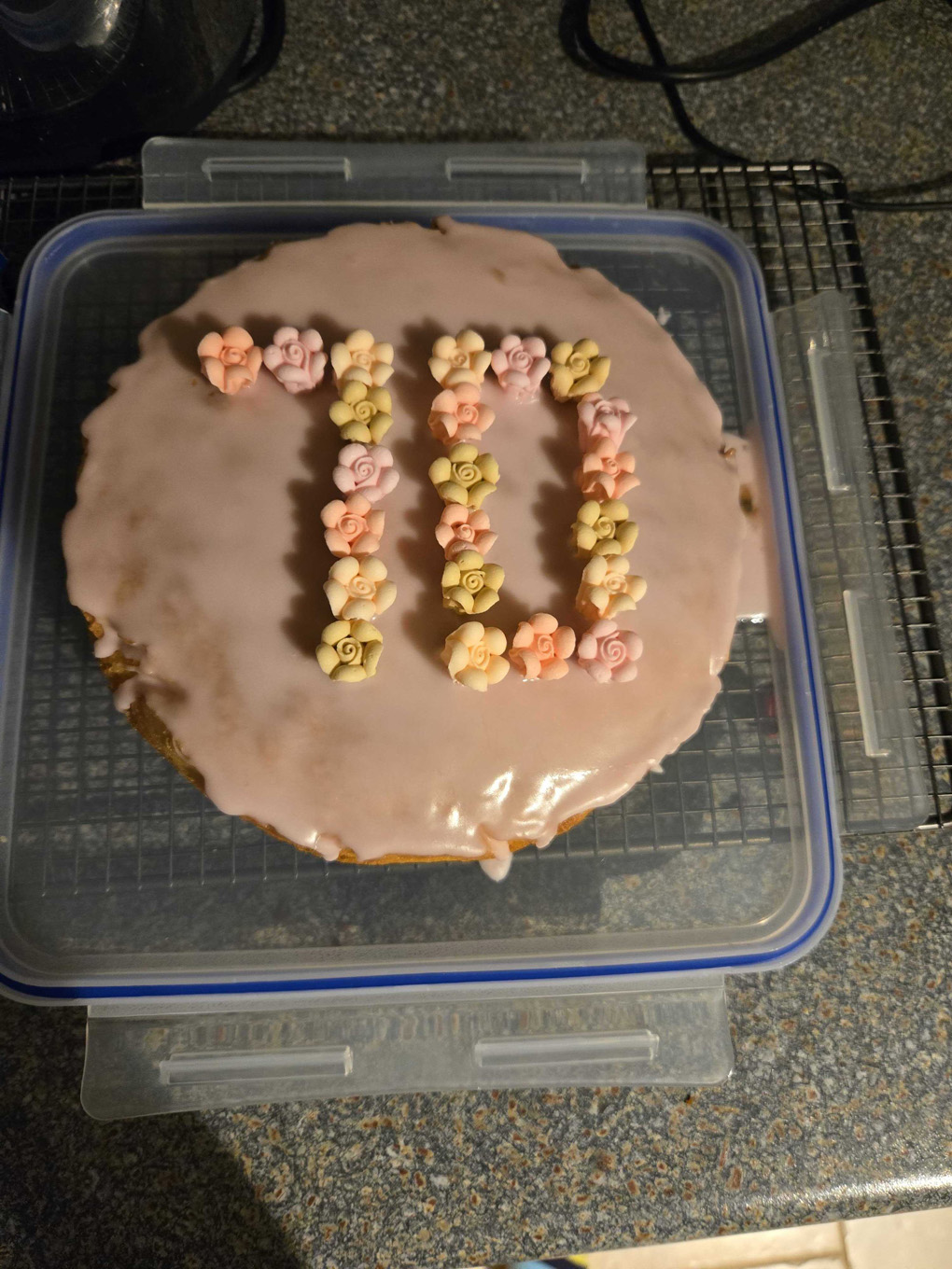 Photo looking directly down on a Victoria sponge cake. It has not fully risen and is not totally circular. The top is covered in pink icing and has small sugar flowers spelling out 70