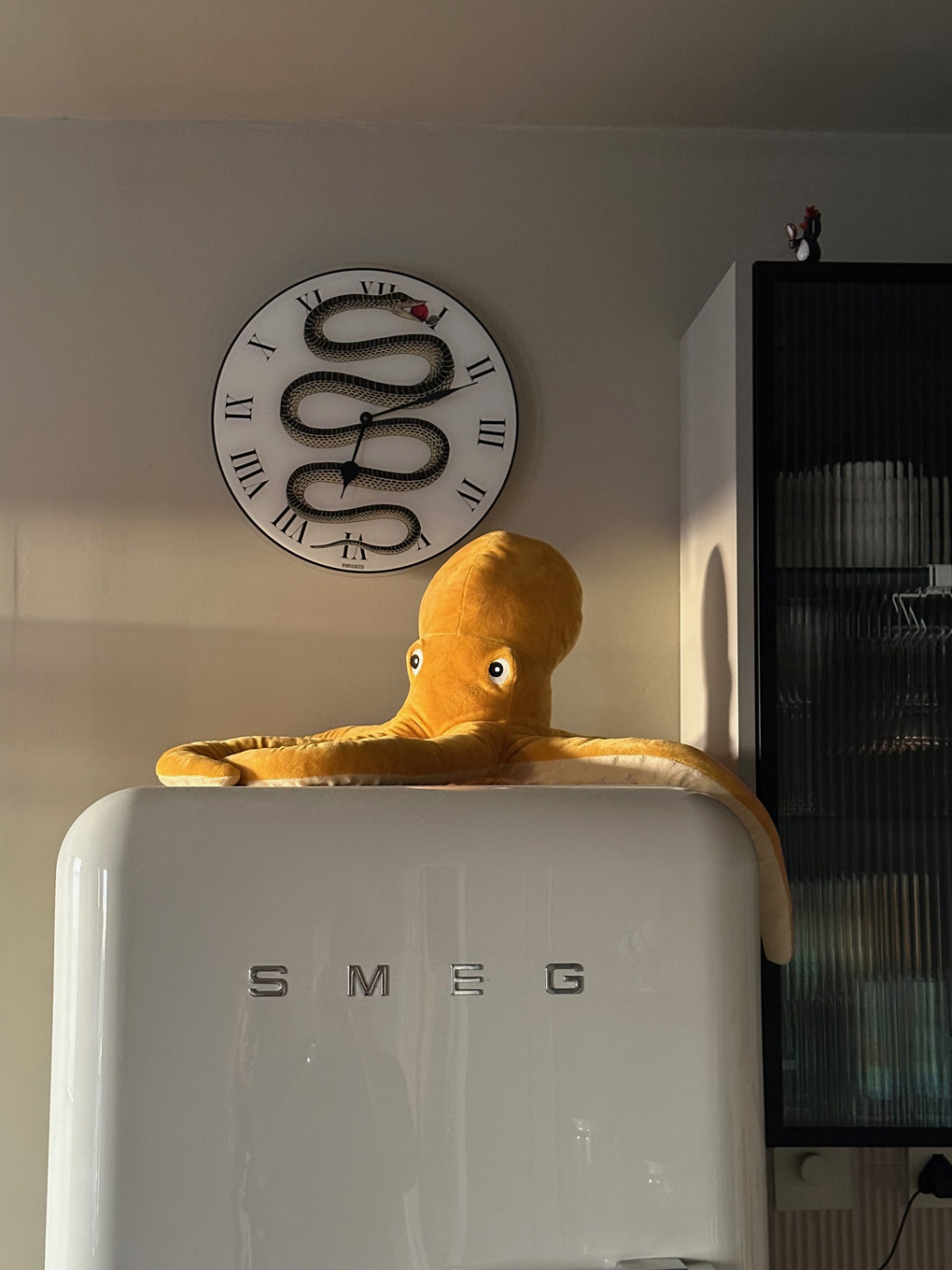A yellow plush octopus sits atop a white SMEG fridge with a clock above it, showing a snake coiled around the clock face with Roman numerals.