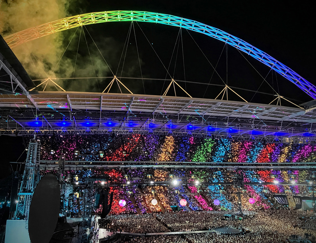A packed Wembley Stadium at night with the crowd wearing glowing wristbands creating rainbow colored patterns across the stands. The iconic stadium arch is lit in rainbow colors with smoke rising into the night sky, and a large stage is visible at the bottom.