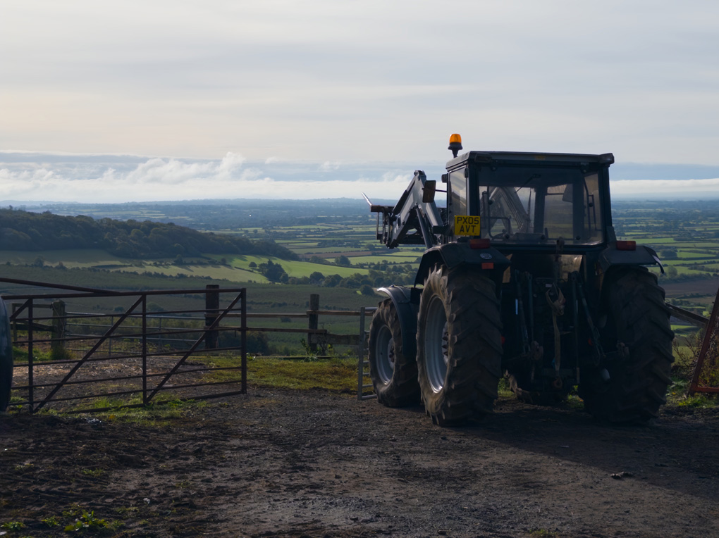 A dark coloured tractor with a bailing fork on the front faces towards a wooded valley with low, bright blue-white clouds in the distance