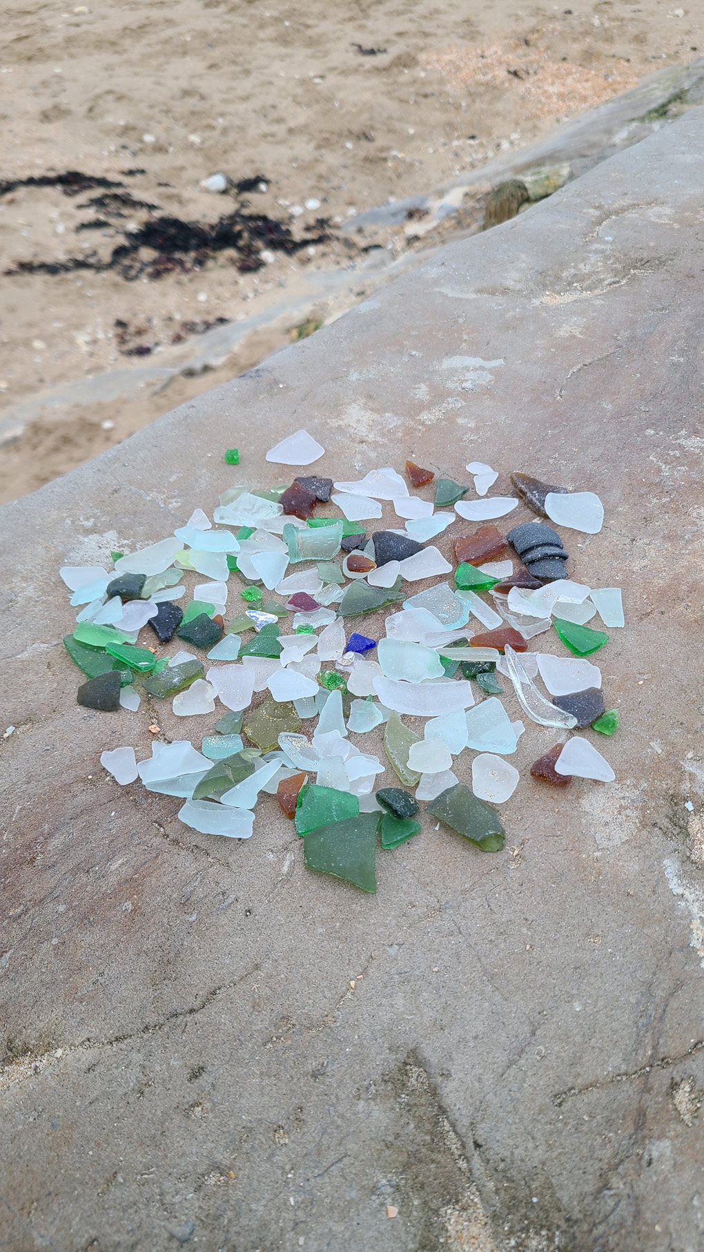 A large pile of sea glass on a concrete barrier by the beach. The glass is mostly frosted whites, greens and browns, with a few blues and reds.