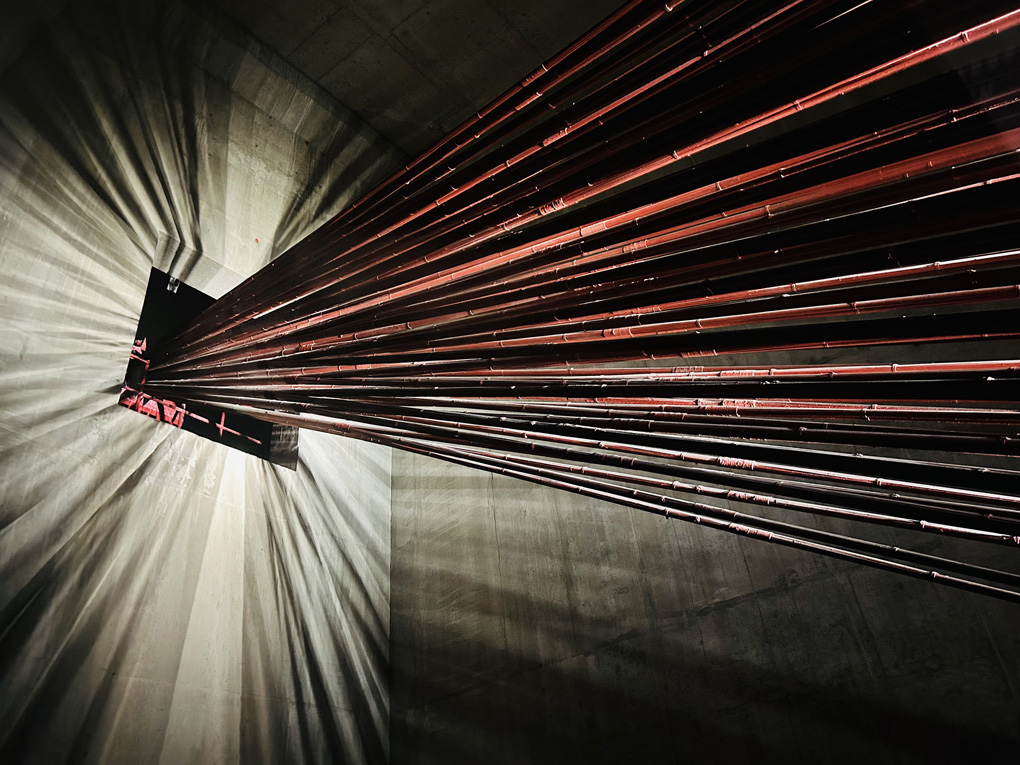 Interior lit concrete room with multiple red steel cables emanating from a square hole. The cables are long and straight, giving of spectral light and reflection their red colouring in the shadows.