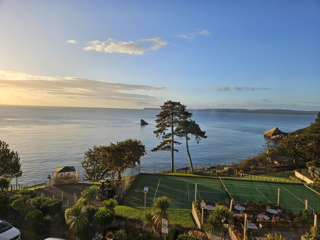 Glorious blue sea meets blue sky with a few whispy clouds. In foreground a tennis court and green trees are enjoying being lit by the sunlight.