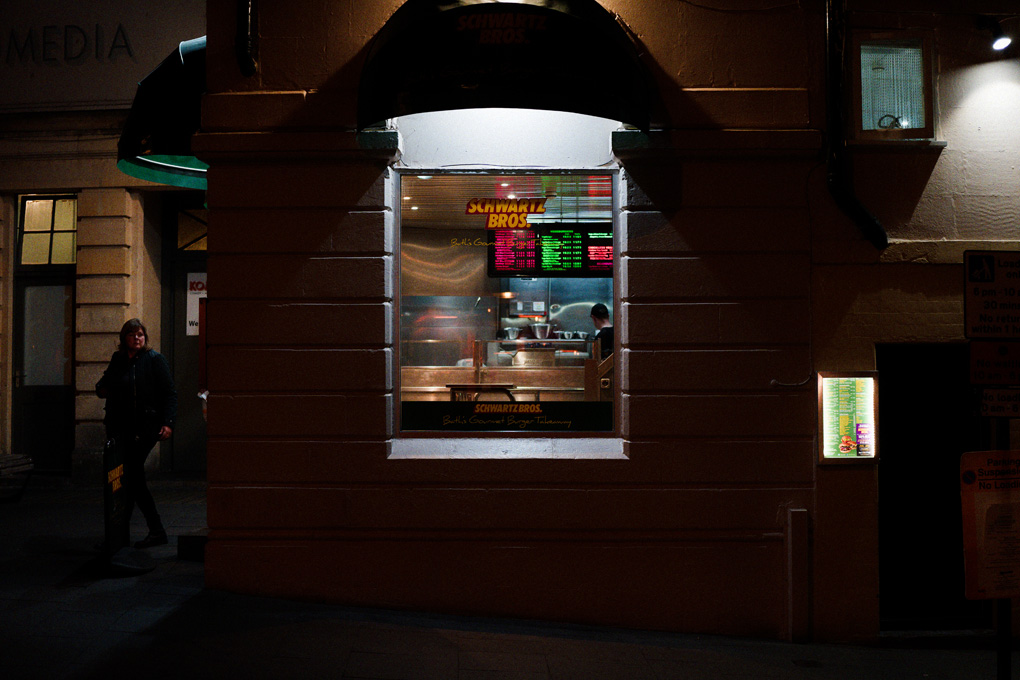 A woman steps out of Schwartz burgers in Bath, on the phone. The burger joint is lit in bright neon contrasting with the sharp shadow lines. It looks cool, 80s, and tasty as fuck.