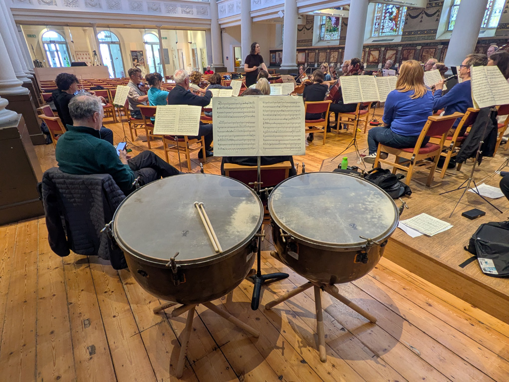 A view from behind two timpani looking out at the rest of the orchestra in a church.