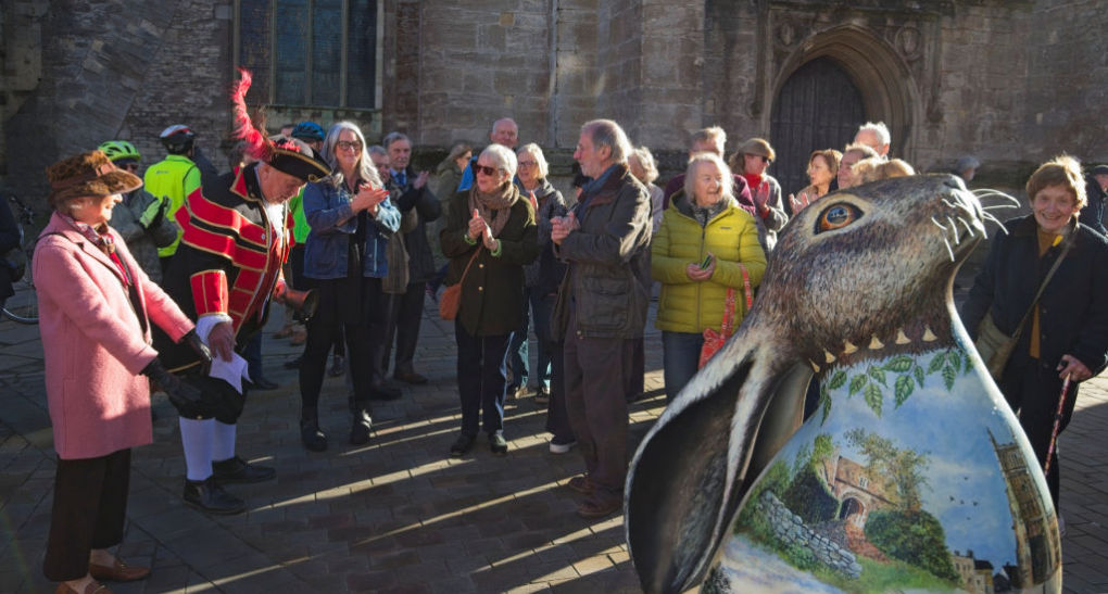 A crowd has gathered for the official welcoming of a new hare statue. The town crier resplendent in his red uniform bows to the artist. The hare in the right foreground is decorated with iconic scenes of both old and new Cirencester