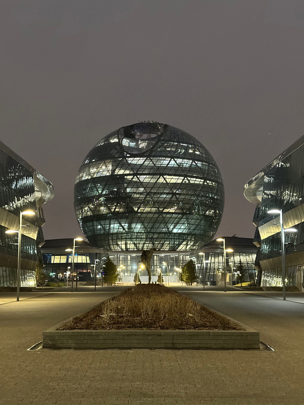 Spherical building at night with lights on inside. Flanked by other buildings on either side.