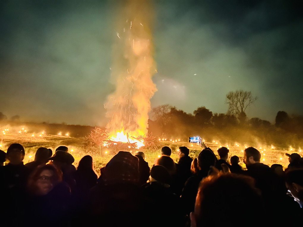 A nighttime bonfire with a column of fire stretching into the night sky. Crowds in the foreground enjoying the glow.