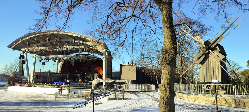 An outdoor stage before the crowd arrive. There's snow on the ground, a large soundsystem, screen and, on the far right, a windmill.