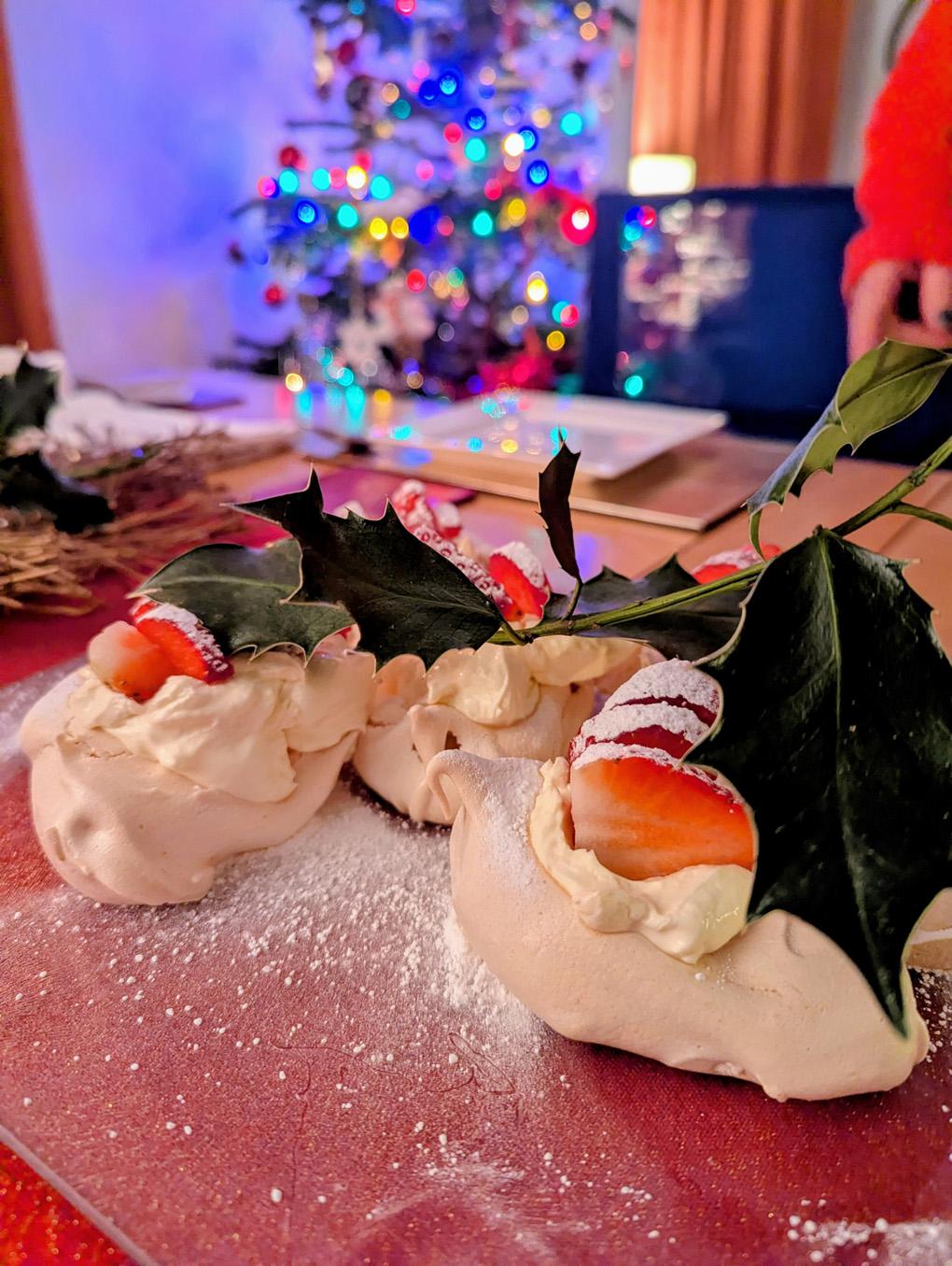 A plate of meringues, cream and strawberries with a sprig of holly on top, against a backdrop of a Christmas tree all lit up