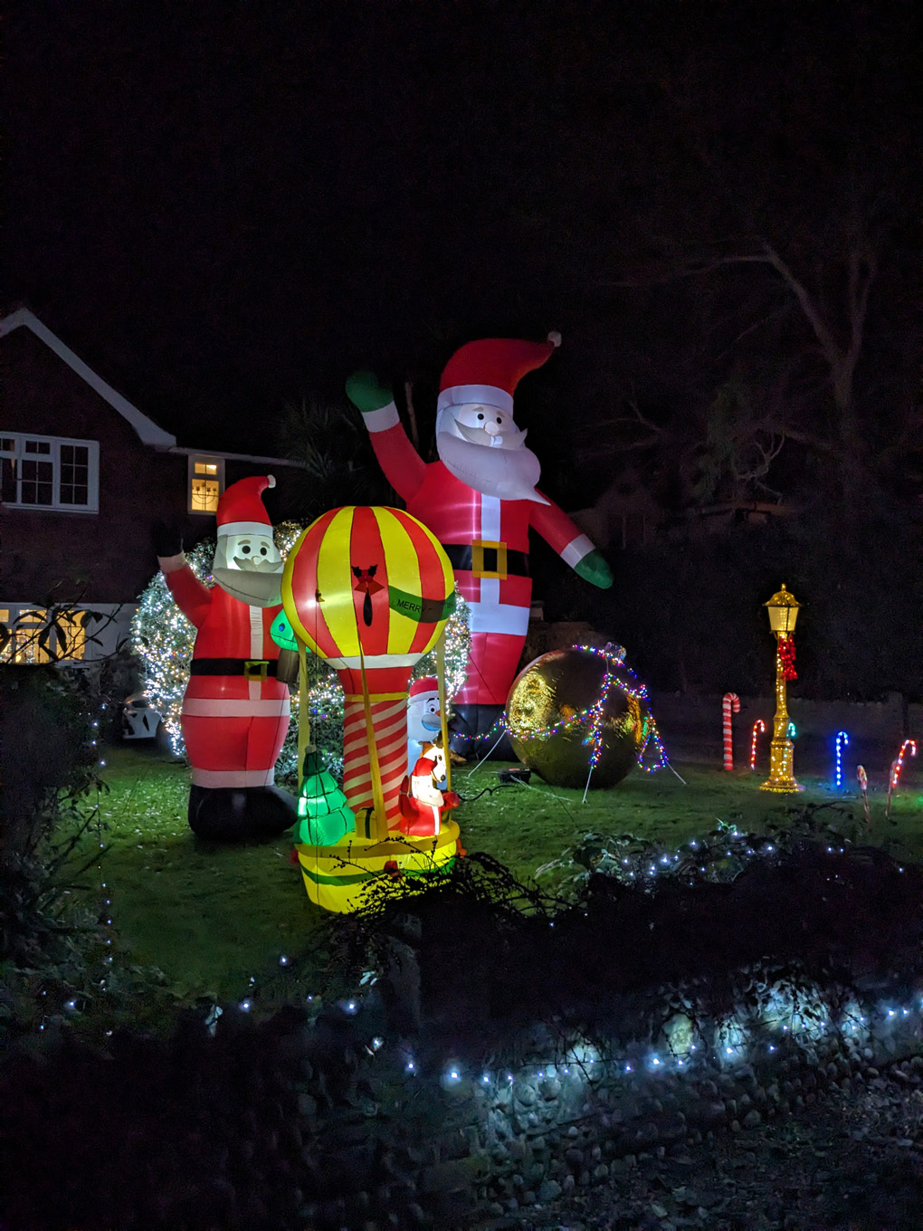 The night is lit up by Christmas decorations in the front garden of a house. Two inflatable Santas, a hot air balloon and a giant bauble that looks a bit like a nautical buoy swathed in fairy lights. One of the inflatable Santas is as tall as the house behind it.