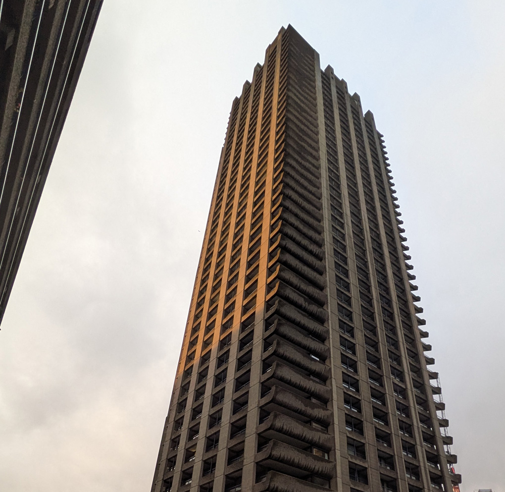 The view from the foot of one of the Barbican Estate towers looking up, its angular concrete floors and balconies disappearing almost to a point with the late low sun catching one side in golden light