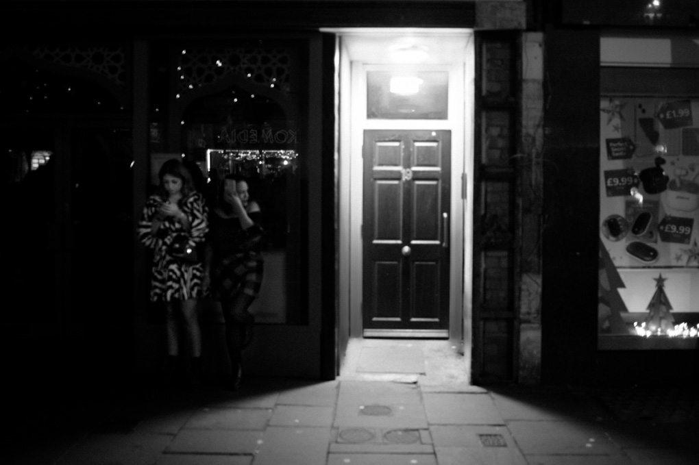 Two women in black and white wait in shadow next to a bright entryway. they are dressed up, probably for a gig at the komedia