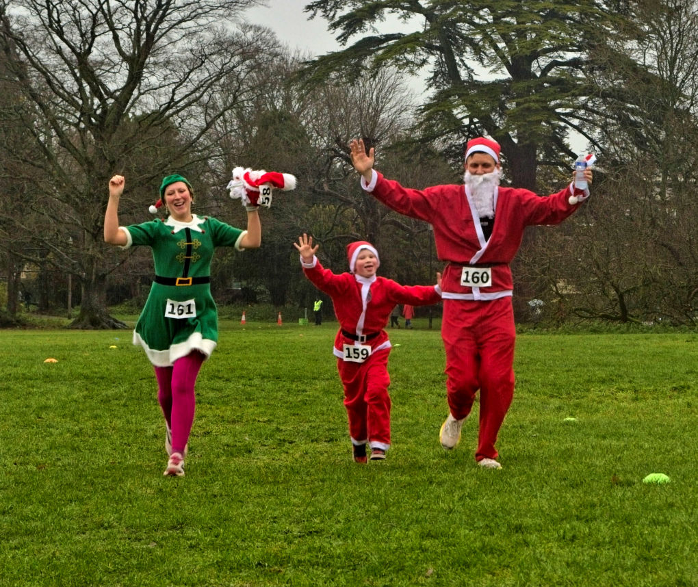 A simple image of a Dad and son dressed as Santas and mum as an elf. They are just finishing the fun run and all hold their arms aloft for the camera.