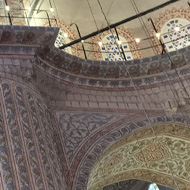 The interior of a mosque - looking upwards towards a dome, balconies and a pillar. The pillar has a tiled patteren on it, depicting rows of foliage - but three of the rows are a significantly deeper shade of blue.