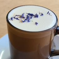 A brown glass mug on a white ceramic saucer. The mug is filled with creamy frothy liquid, topped with pretty purple and blue flower petals. The mugs handle is facing to the right, there is a silver teaspoon on the saucer under the handle. It's on a round, light wooden table.