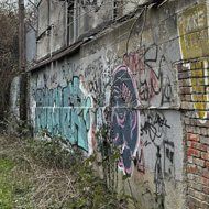 A partly crumbling brick and concrete building, covered in brightly coloured graffiti tags, which stands next to a long, curving tarmac pathway
