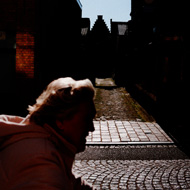 A woman rides a bicycle past a sunlit alley on a cobbled street in Ypres. Sunlight glances off her hair and jacket, and some geometric shapes in the background.