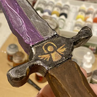 A model knife with a purple and silver blade, and a stylised angel on the hilt, being held in front of some model painting supplies.