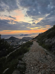 The foreground shows a very rocky path going down a steep hill. The background shows the sun setting on a sea with a few small islands